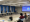 A man speaks to a small group in a conference room with a slide showing his name and title.
