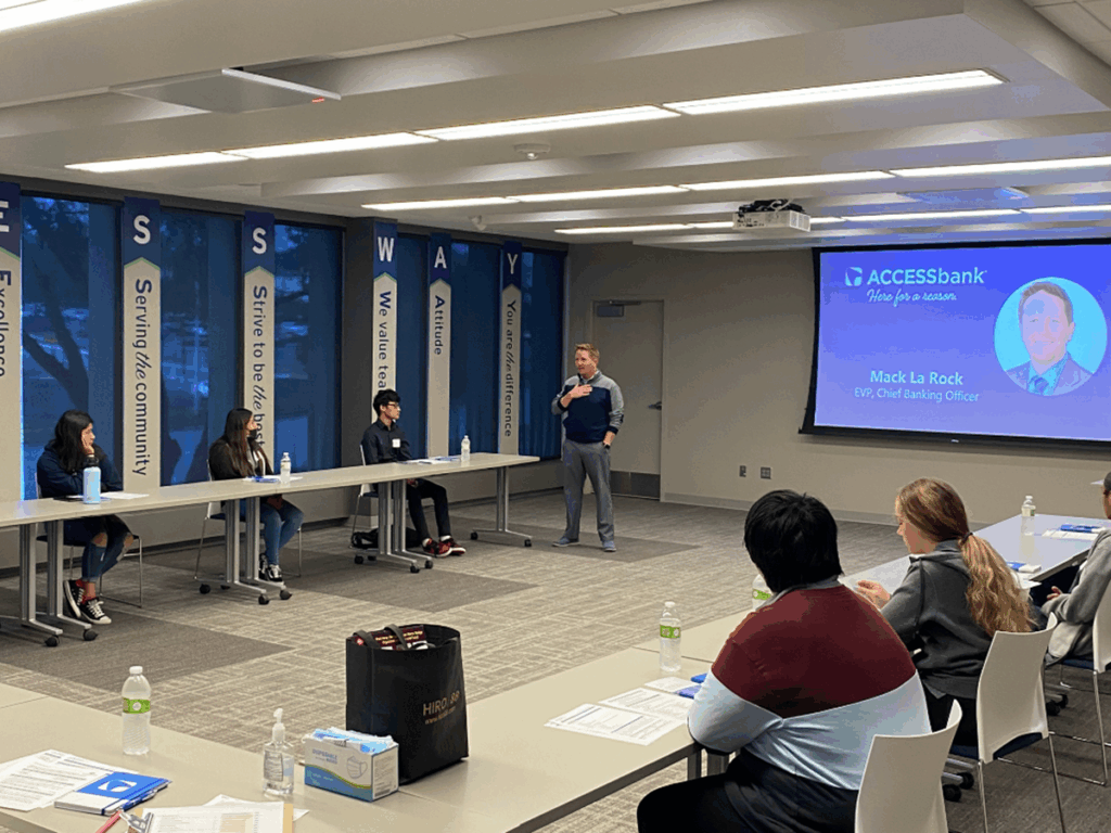 A man speaks to a small group in a conference room with a slide showing his name and title.
