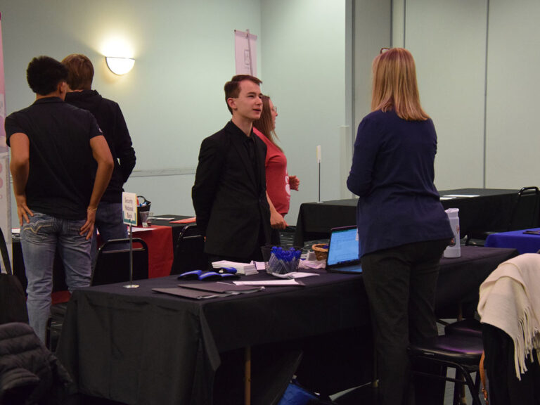 A young man in a black suit appears confident while speaking with a woman in a blue cardigan.