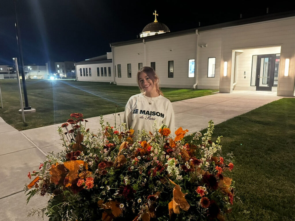 Tala Serhan stands behind a large bundle of floral arrangements. The scene is at night in front of a lit building.