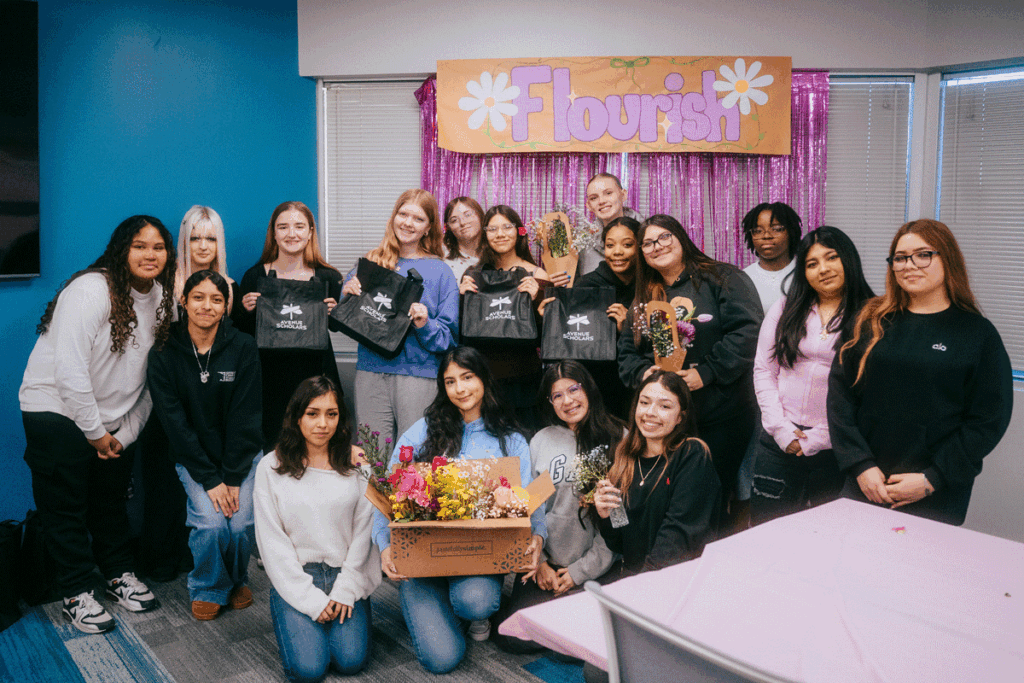 Group of high school students pose together indoors beneath a “Flourish” banner, holding Avenue Scholars bags and small floral arrangements; one student kneels in front holding a box of flowers, while others stand behind smiling.