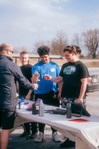 A small group of high school students stands around a table outdoors, smiling as an adult demonstrates a hands-on activity with automotive fluids and tools.