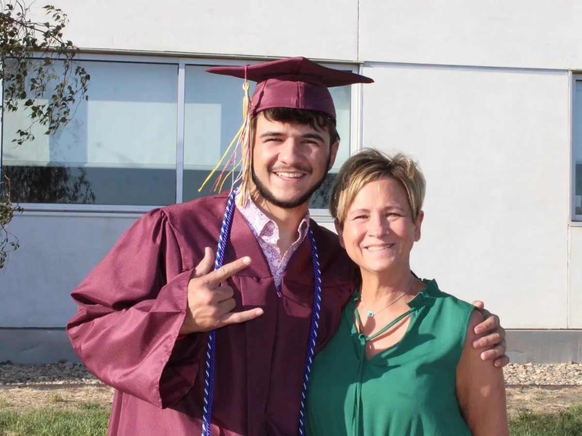 a high school graduate in cap and gown stands next to a woman in celebratory fashion.