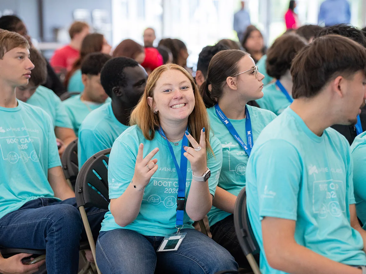 An Avenue Scholars Boot Camp participant smiles and flashes a peace sign while seated with fellow Scholars during a group session at the 2026 graduation.